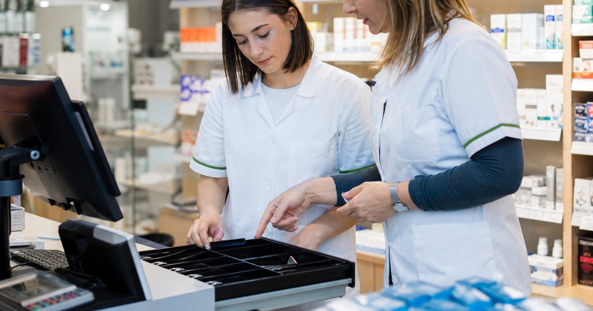 A young trainee cashier and an experienced coworker standing together behind a checkout counter in a small pharmacy, the coworker pointing into the open cash drawer, bright retail lighting